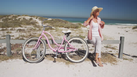 Ältere-Kaukasische-Frau,-Die-Auf-Einer-Barriere-Neben-Einem-Fahrrad-Am-Strand-Sitzt