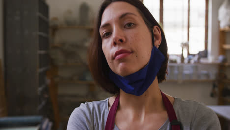 Portrait-of-female-potter-with-lowered-face-mask-at-pottery-studio