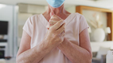 Mid-section-of-senior-caucasian-woman-wearing-face-mask-sanitizing-her-hands-at-home