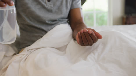 Mid-section-of-african-american-senior-man-sanitizing-his-hands-while-sitting-on-the-bed-at-home