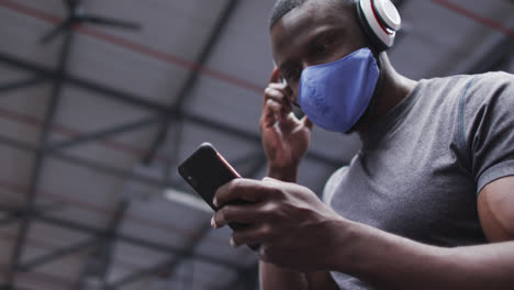 Hombre-Afroamericano-Con-Mascarilla-Usando-Un-Teléfono-Inteligente-En-El-Gimnasio