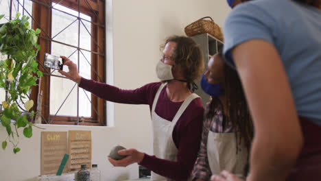 Diverse-potters-wearing-face-masks-and-aprons-holding-clay-taking-a-selfie-using-smartphone-at-potte