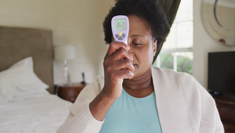 African-american-senior-woman-measuring-her-temperature-while-sitting-on-the-bed-at-home