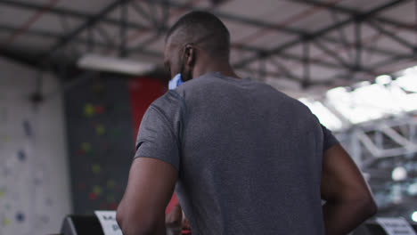 Fit-african-american-man-wearing-face-mask-running-on-treadmill-in-the-gym