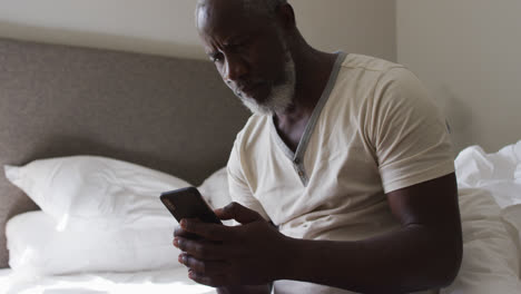 Afroamerikanischer-Senior,-Der-Sein-Smartphone-Benutzt,-Während-Er-Zu-Hause-Auf-Dem-Bett-Sitzt