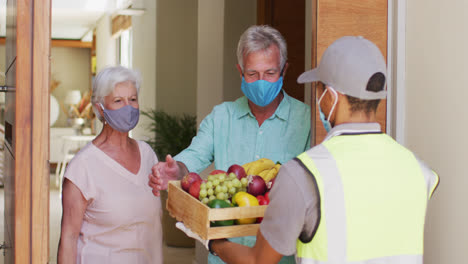 Delivery-man-delivering-groceries-to-senior-caucasian-couple-wearing-face-masks-at-home