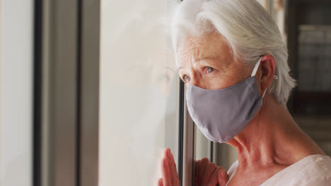 Mujer-Caucásica-Mayor-Con-Mascarilla-Mirando-Por-La-Ventana-En-Casa