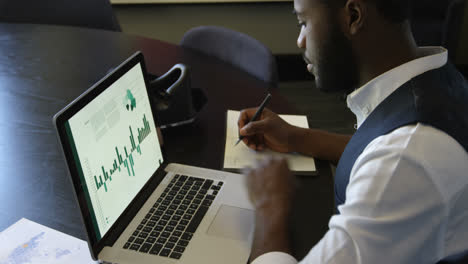 Side-view-of-young-black-businessman-writing-on-diary-while-using-working-on-laptop-in-office-4k