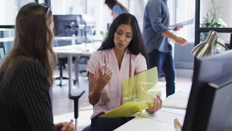 Mixed-race-businesswoman-holding-file-and-talking-to-female-colleague-at-casual-office-meeting