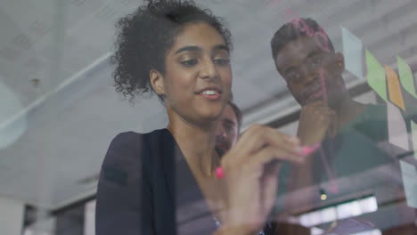 Diverse-colleagues-using-memo-notes-and-writing-on-glass-wall-having-a-discussion