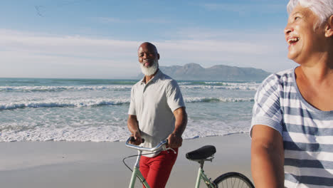 Smiling-senior-african-american-couple-walking-with-bicycles-at-the-beach