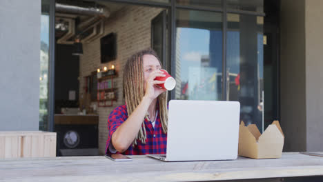 Hombre-De-Raza-Mixta-Con-Rastas-Sentado-En-La-Mesa-Afuera-De-La-Cafetería-Tomando-Café-Y-Usando-Una-Computadora-Portátil