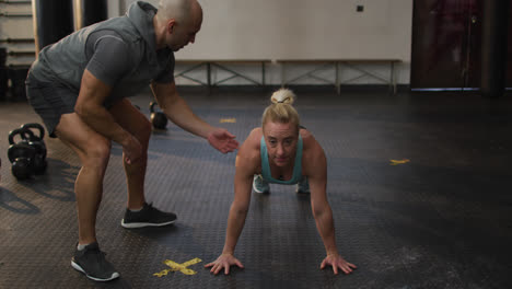 Male-trainer-correcting-form-of-fit-caucasian-woman-while-push-up-exercise-at-the-gym