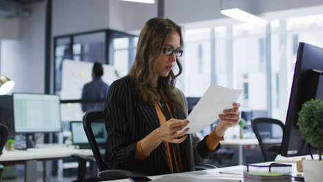 Mujer-De-Negocios-Caucásica-Sentada-En-El-Escritorio-Leyendo-Un-Documento-Y-Quitándose-Las-Gafas-En-La-Oficina