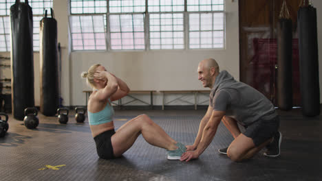 Colocar-A-Una-Mujer-Caucásica-Realizando-Abdominales-Mientras-Un-Entrenador-Masculino-Sostiene-Sus-Piernas-En-El-Gimnasio.