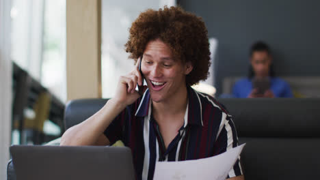 Caucasian-man-going-through-paperwork-talking-on-smartphone-using-laptop-in-the-office