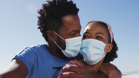 Portrait-of-african-american-couple-wearing-face-masks-looking-to-camera-on-the-beach