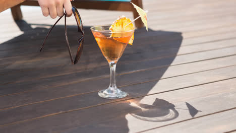 Summer-cocktail-by-person-holding-sunglasses-in-sun-lounger-and-swimming-pool