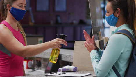 Two-caucasian-women-wearing-face-masks-talking-at-reception-of-gym-and-disinfecting-hands