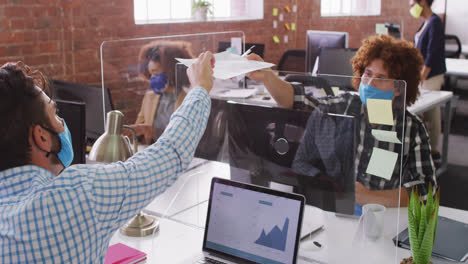 Diverse-business-colleagues-exchanging-documents-over-sneeze-screen-wearing-face-masks