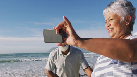 Smiling-senior-african-american-couple-walking-with-bicycles-and-taking-a-selfie-at-the-beach