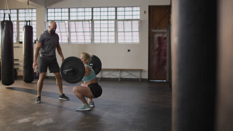 Caucasian-fit-woman-wearing-face-mask-and-lifting-weight-bar-with-coach