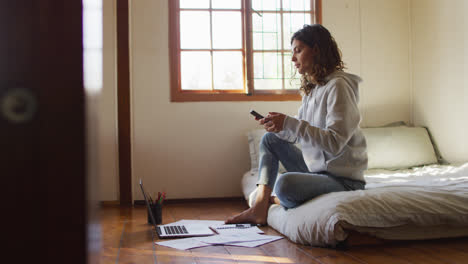 Mixed-race-woman-working-at-home,-sitting-on-bed-using-smartphone-and-laptop-in-cottage