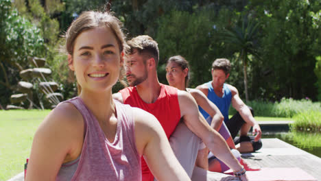 Lächelnde-Kaukasische-Frau-Praktiziert-Yoga-Mit-Einer-Vielfältigen-Gruppe,-Die-Im-Sonnigen-Park-Sitzt