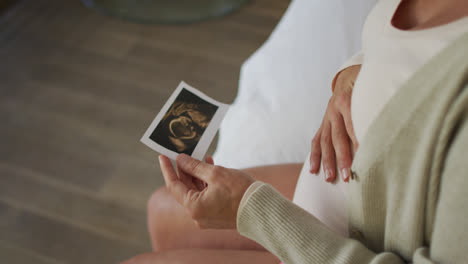 Manos-De-Una-Mujer-Embarazada-Caucásica-Sentada-En-La-Cama,-Tocando-El-Vientre-Y-Mirando-La-Foto-De-Ustrasound