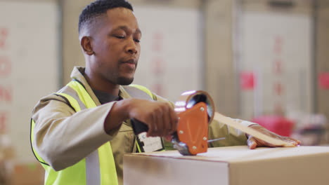 African-american-male-worker-wearing-safety-suit-and-packing-boxes-in-warehouse