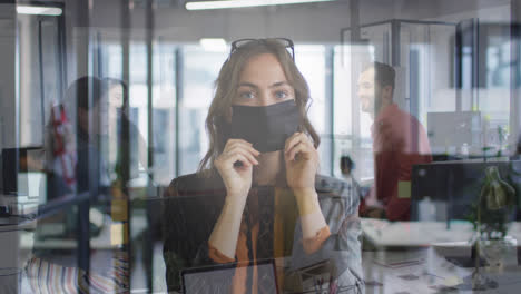 Composite-of-caucasian-woman-wearing-face-mask-in-office,-and-colleagues-talking