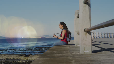 Animation-of-light-spots-over-biracial-woman-sitting-with-binoculars-on-promenade