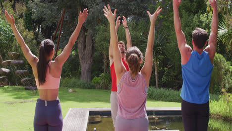 Vielfältige-Gruppe-Und-Männlicher-Lehrer-Praktizieren-Yoga,-Stehen-Und-Heben-Die-Arme-Im-Sonnigen-Park