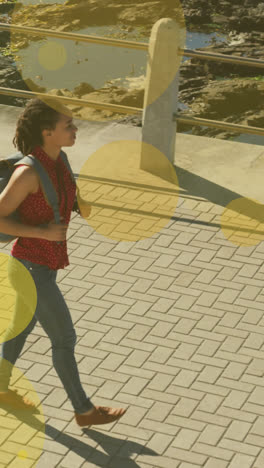 Animation-of-yellow-spots-over-biracial-woman-walking-on-promenade