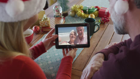 Caucasian-couple-with-santa-hats-using-tablet-for-christmas-video-call-with-couple-on-screen