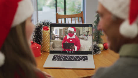 Vista-Trasera-De-Una-Pareja-Caucásica-Con-Gorros-De-Papá-Noel-Teniendo-Una-Videollamada-En-Una-Computadora-Portátil-Durante-La-Navidad