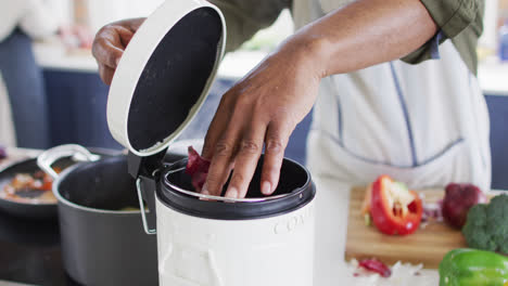 Mid-section-of-african-american-senior-man-wearing-apron-putting-vegetable-peelings-in-a-compost-bin