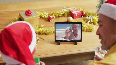 Kaukasischer-Vater-Und-Sohn-Mit-Weihnachtsmützen-Nutzen-Tablet-Für-Weihnachtsvideoanruf-Mit-Paar-Auf-Dem-Bildschirm