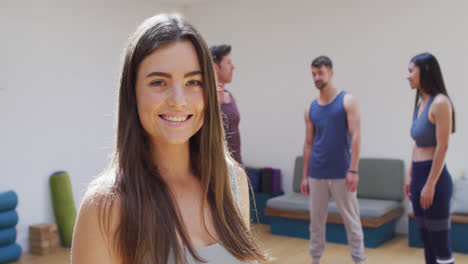 Retrato-De-Una-Mujer-Caucásica-Sonriente,-Con-Un-Grupo-Diverso-En-Clase-De-Yoga-Hablando-En-Segundo-Plano