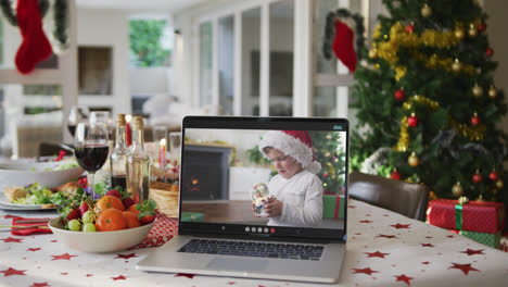 Glücklicher-Kaukasischer-Junge-Mit-Weihnachtsmütze-Auf-Dem-Laptop,-Der-Auf-Dem-Weihnachtstisch-Liegt