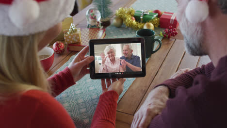 Kaukasisches-Paar-Mit-Weihnachtsmützen-Nutzt-Tablet-Für-Weihnachtsvideoanruf-Mit-Paar-Auf-Dem-Bildschirm