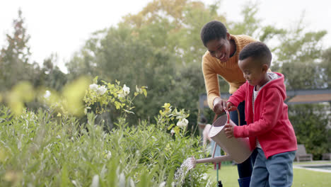 Feliz-Hijo-Afroamericano-Y-Madre-Regando-Plantas-En-Un-Jardín-Soleado,-Cámara-Lenta