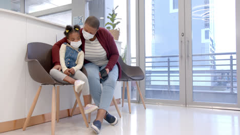 African-american-mother-and-daughter-wearing-face-masks-holding-smartphone-in-hospital,-slow-motion