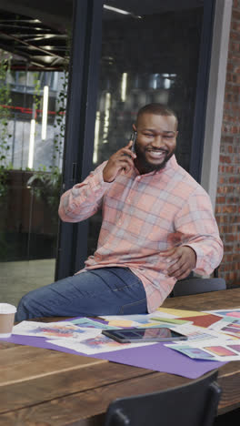Vertical-video-of-happy-african-american-casual-businessman-sitting-on-office-desk-talking-on-phone