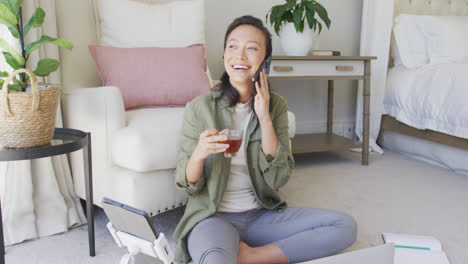 Happy-asian-woman-talking-at-smartphone-and-drinking-tea-in-bedroom,-in-slow-motion