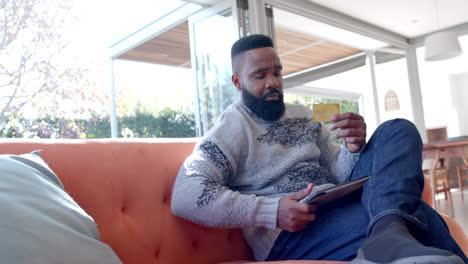 African-american-man-using-tablet-and-credit-card-in-sunny-living-room,-slow-motion