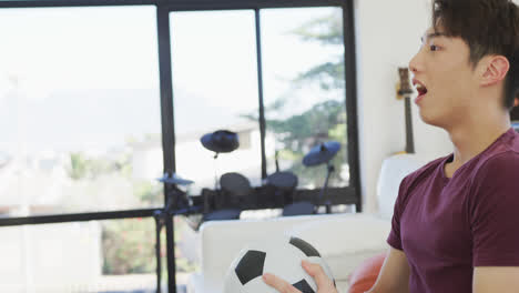 Happy-asian-male-teenager-watching-tv-with-football-and-sitting-in-living-room