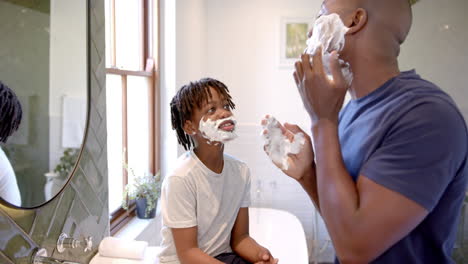 Happy-african-american-father-and-son-applying-shaving-foam-on-face-in-bathroom-at-home,-slow-motion