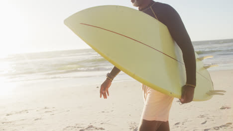 Glücklicher-älterer-Afroamerikanischer-Mann,-Der-In-Zeitlupe-Am-Strand-Spazieren-Geht-Und-Ein-Surfbrett-Hält