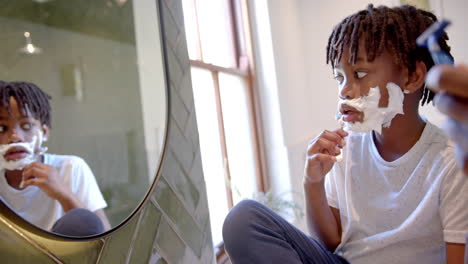 Happy-african-american-father-and-son-shaving-beard-in-bathroom-at-home,-slow-motion